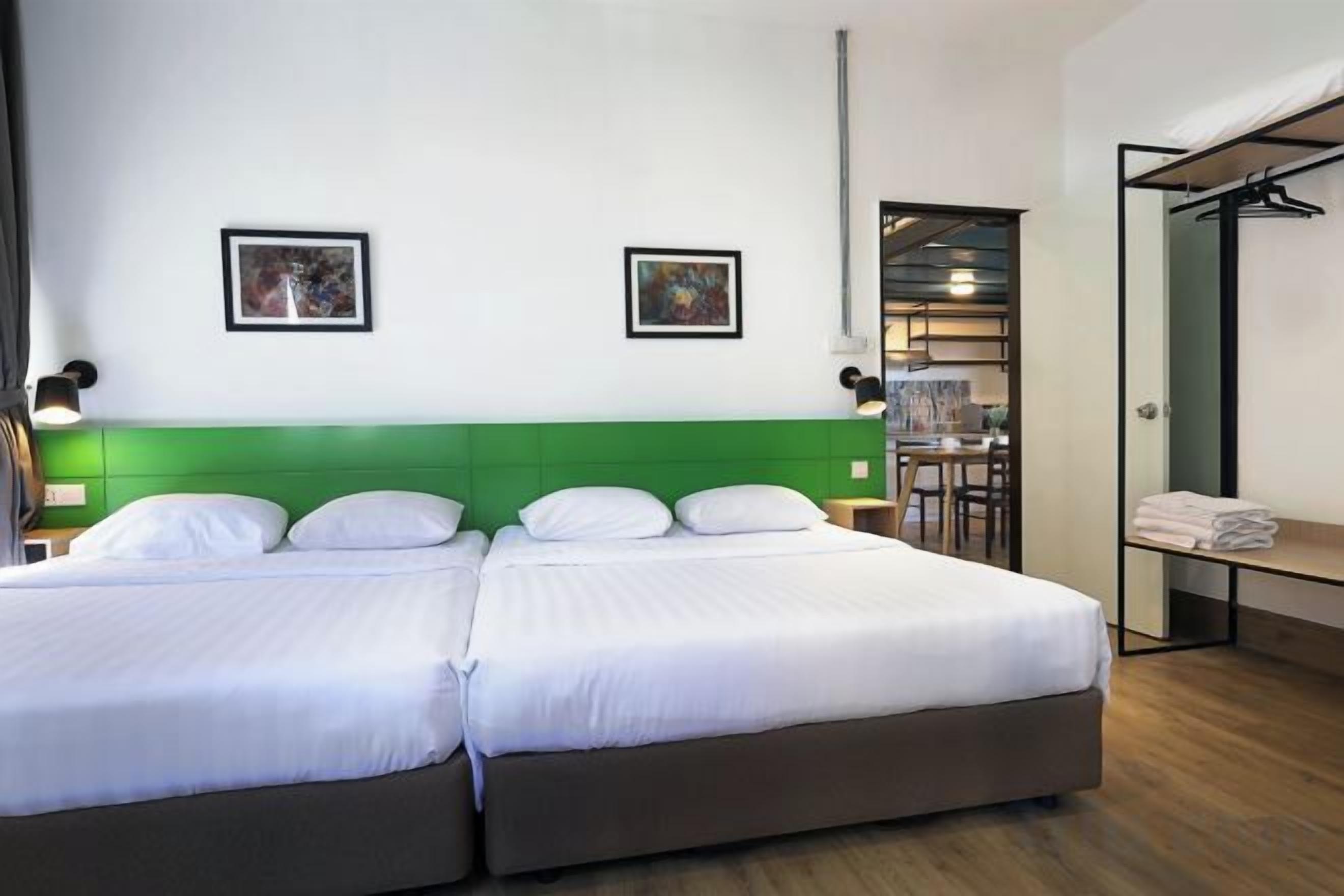 A hotel room with two beds, a green headboard, a desk with a chair, and a coat rack.