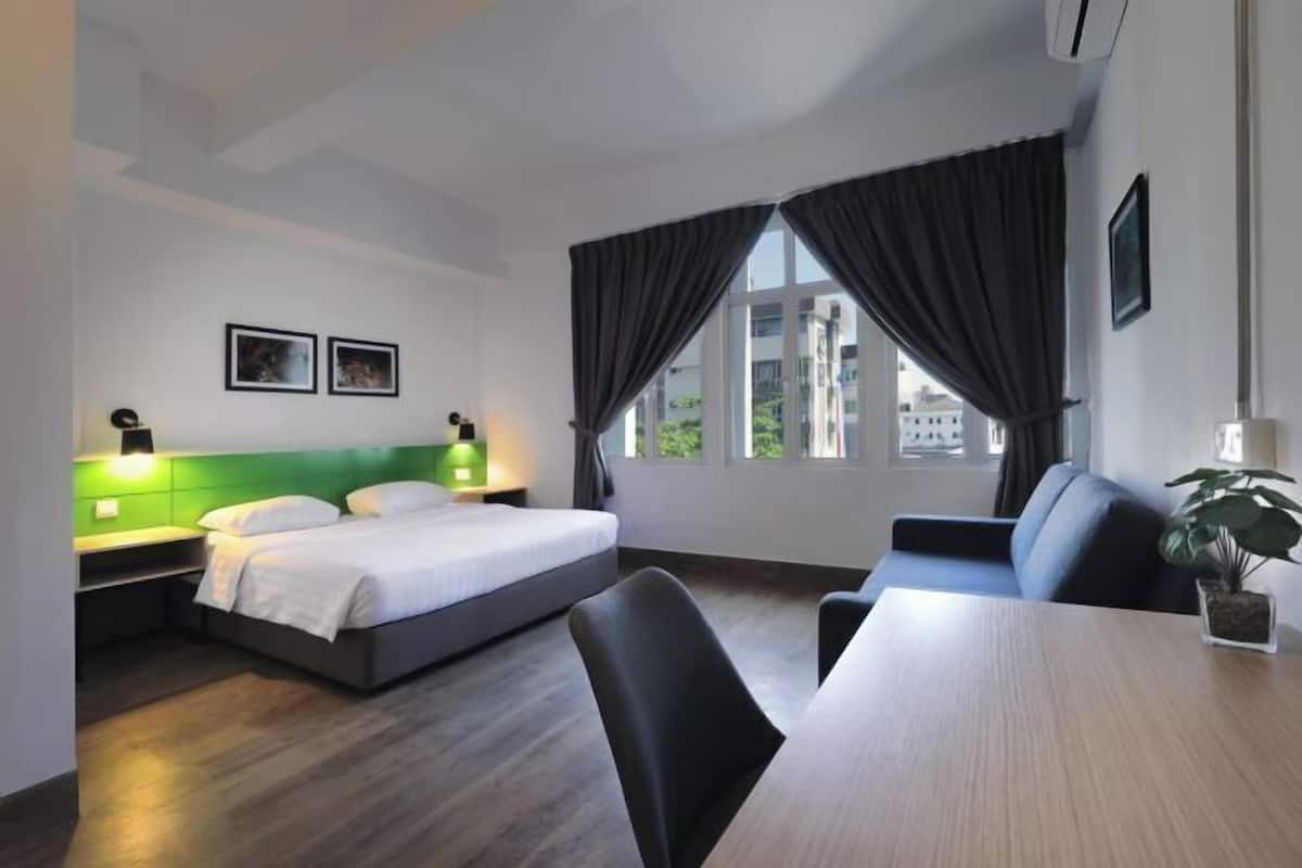 A hotel room with a bed, a desk, and a chair. There are two bedside tables with lamps, a window with curtains, and a plant on the desk.
