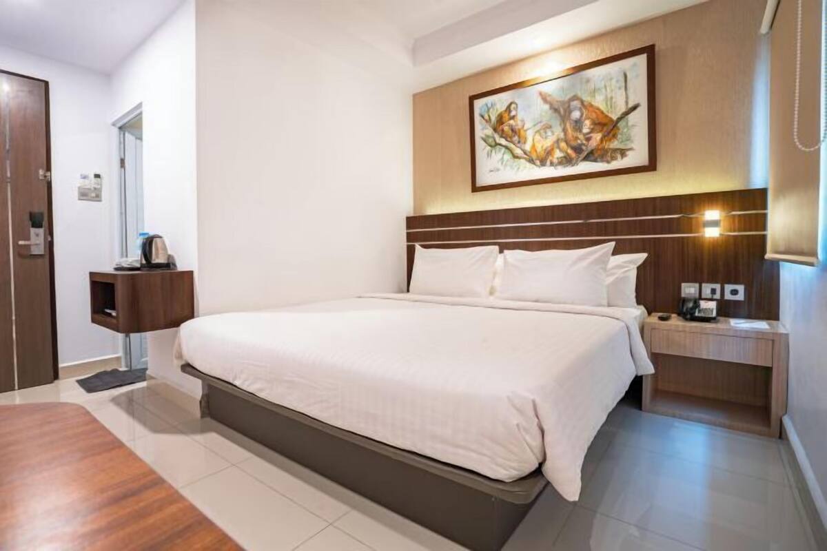 A hotel room with a large bed, a nightstand, a desk, a painting on the wall, and a door.