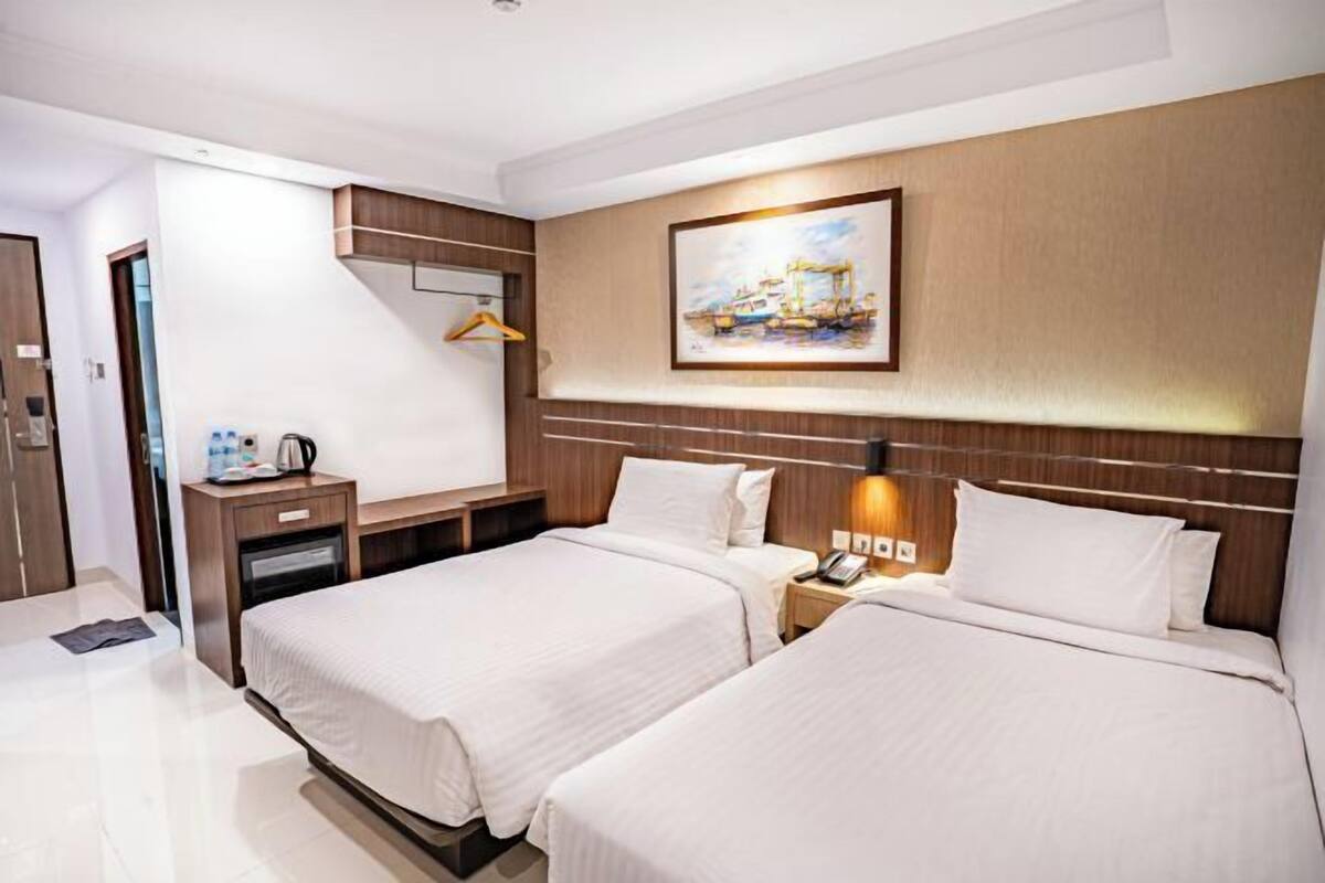 A hotel room with two beds, a wooden headboard, a painting, a desk with a telephone, and a hanging coat rack.