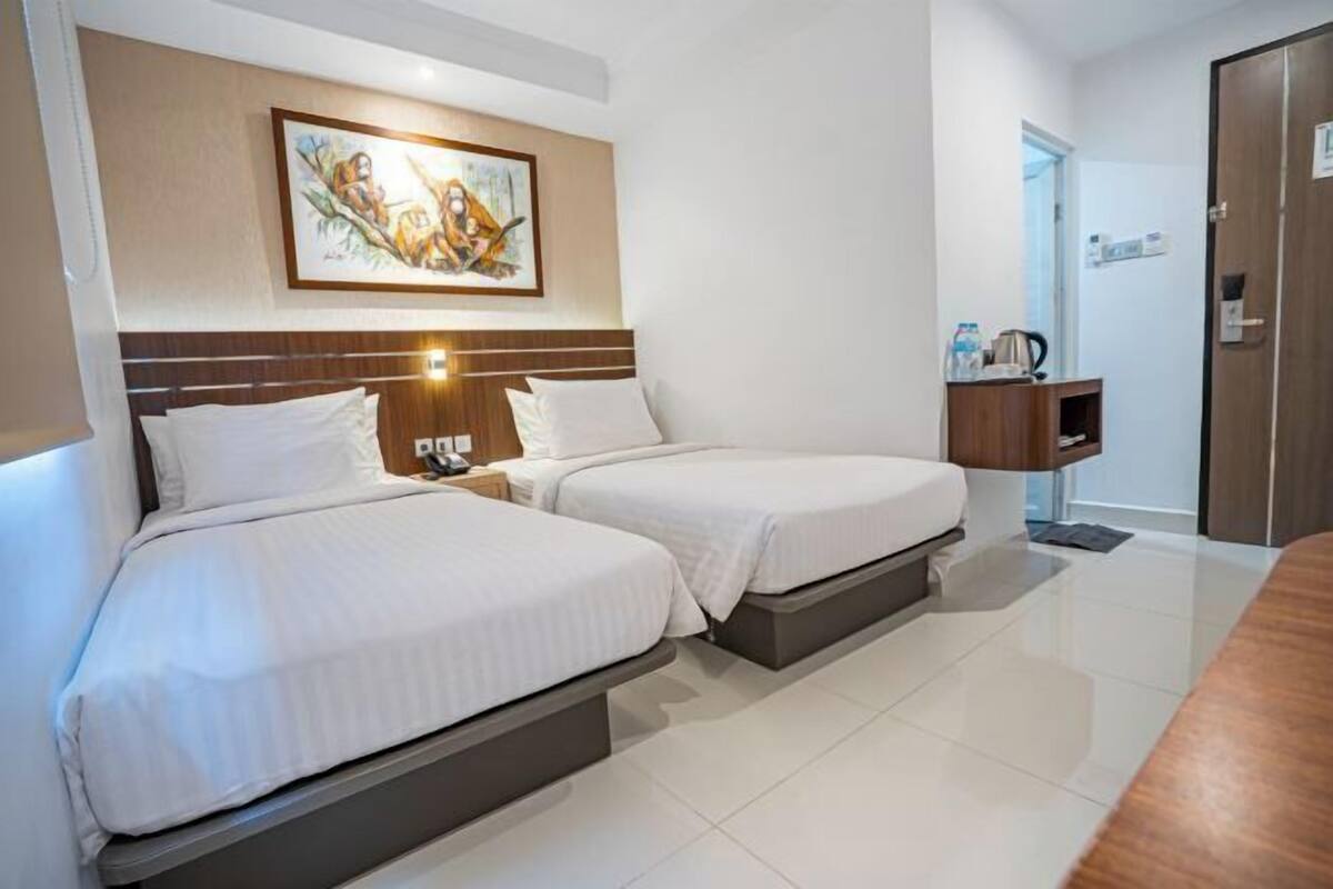 A hotel room with two beds, a wooden headboard, a nightstand, a painting, a small cabinet, and a door.