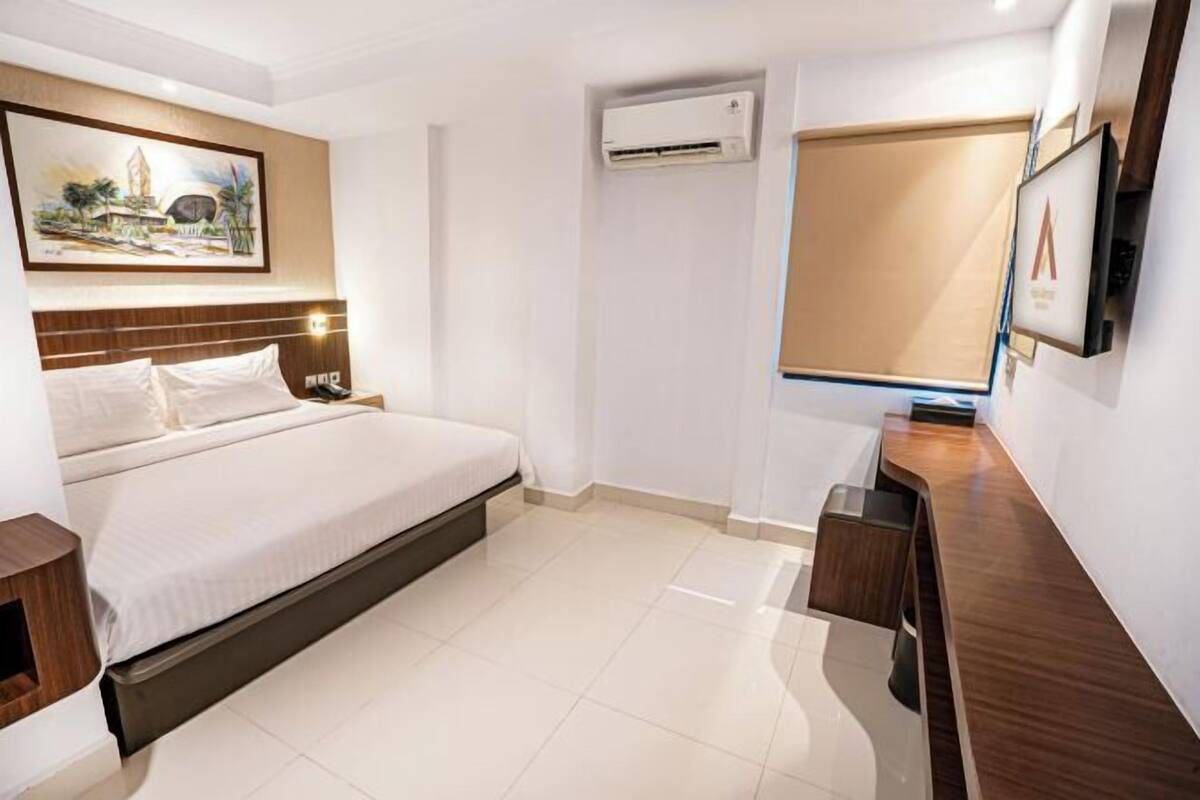 A modern hotel room with a bed, a desk, an air conditioning unit, and a painting on the wall.