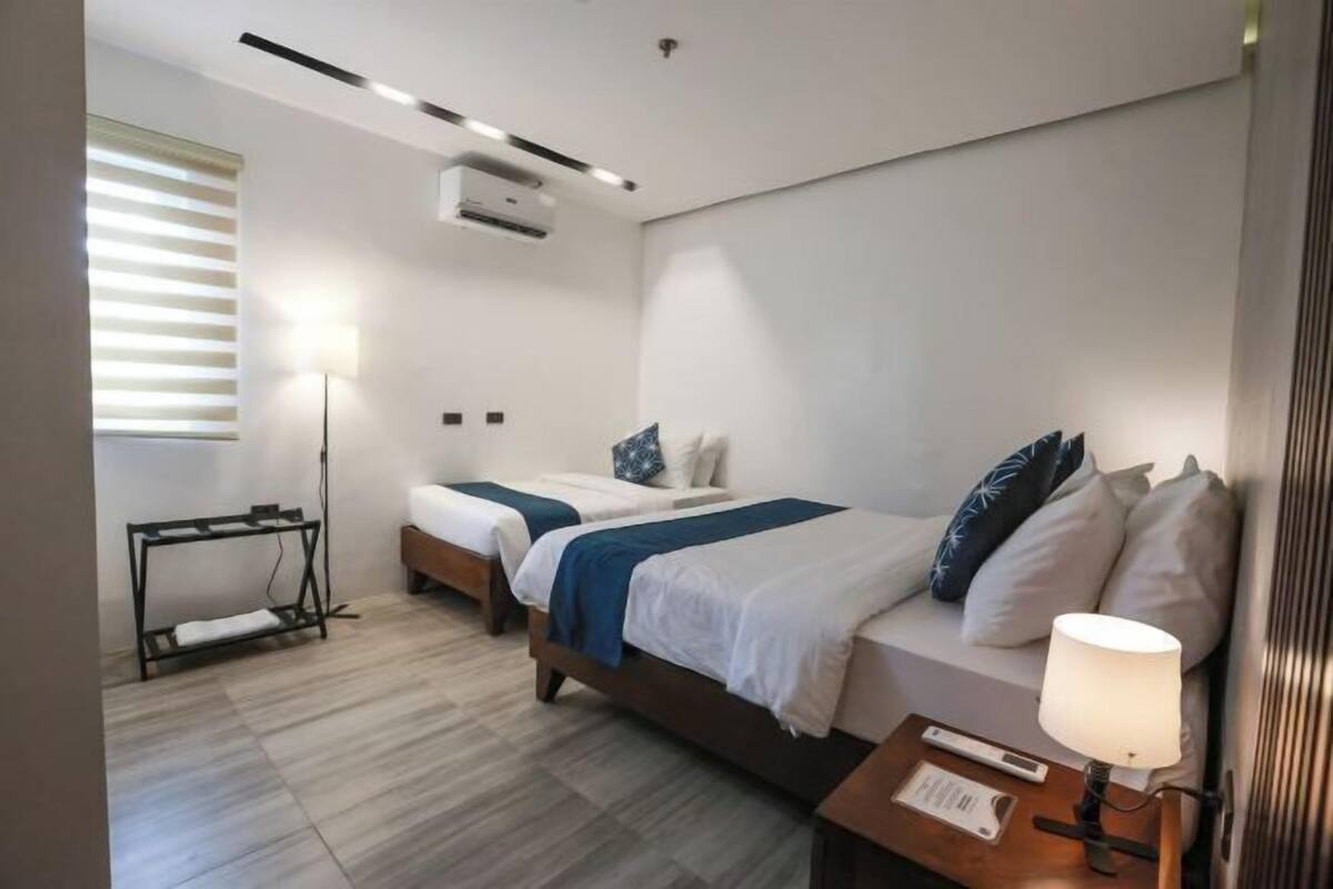 A hotel room with two beds, a standing lamp, a bedside table, and a window with blinds.