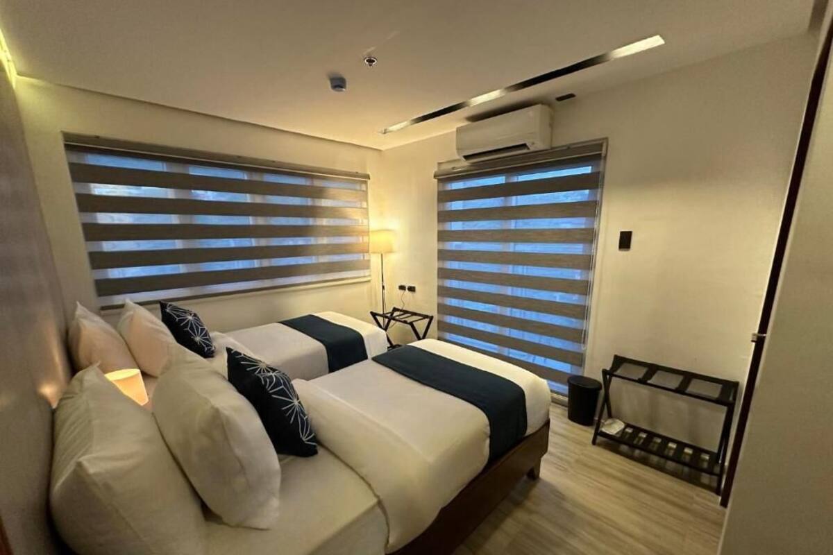 A modern hotel room with two beds, a small seating area, and a window with blinds.