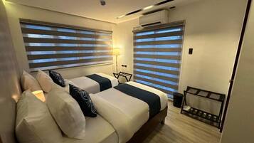A modern hotel room with two beds, a small seating area, and a window with blinds.