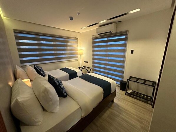 A modern hotel room with two beds, a small seating area, and a window with blinds. - Coron 180 Hotel (Coron)
