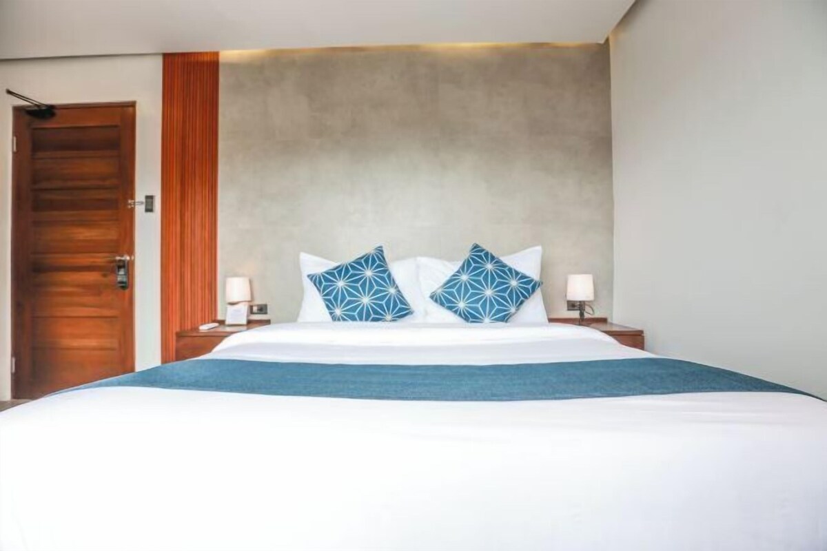 Habitación de hotel con una cama, dos mesitas de noche, una puerta de madera y un cabecero.