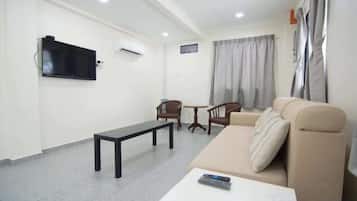 Una sala de estar con un sofá, una mesita, un banco, un televisor montado en la pared y una ventana con cortinas.