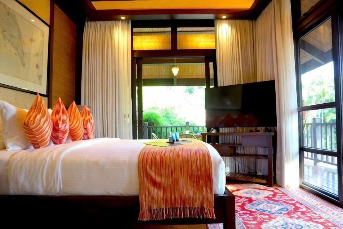 A bedroom with a bed, pillows, a TV, a bookshelf, and a large window.