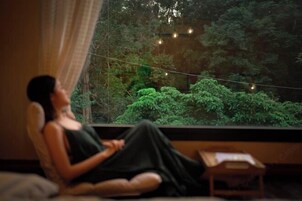 Uma pessoa sentada perto de uma janela com vista para vegetação e luzes de Natal.