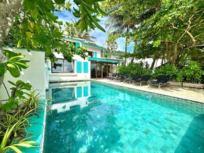Una piscina con agua azul cristalina, rodeada de exuberante vegetación y un edificio con fachada blanca y detalles azul turquesa.