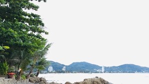 A beach with a rocky outcrop, palm trees, and a distant cityscape. - Villa Goodwill Beach House 3bedrooms on Beach front & private pool Patong Phuket (Tambon Patong)