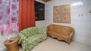 Une pièce comprenant un lit, une chaise en osier, une petite table, une fenêtre avec des rideaux et un carreau décoratif sur le mur.