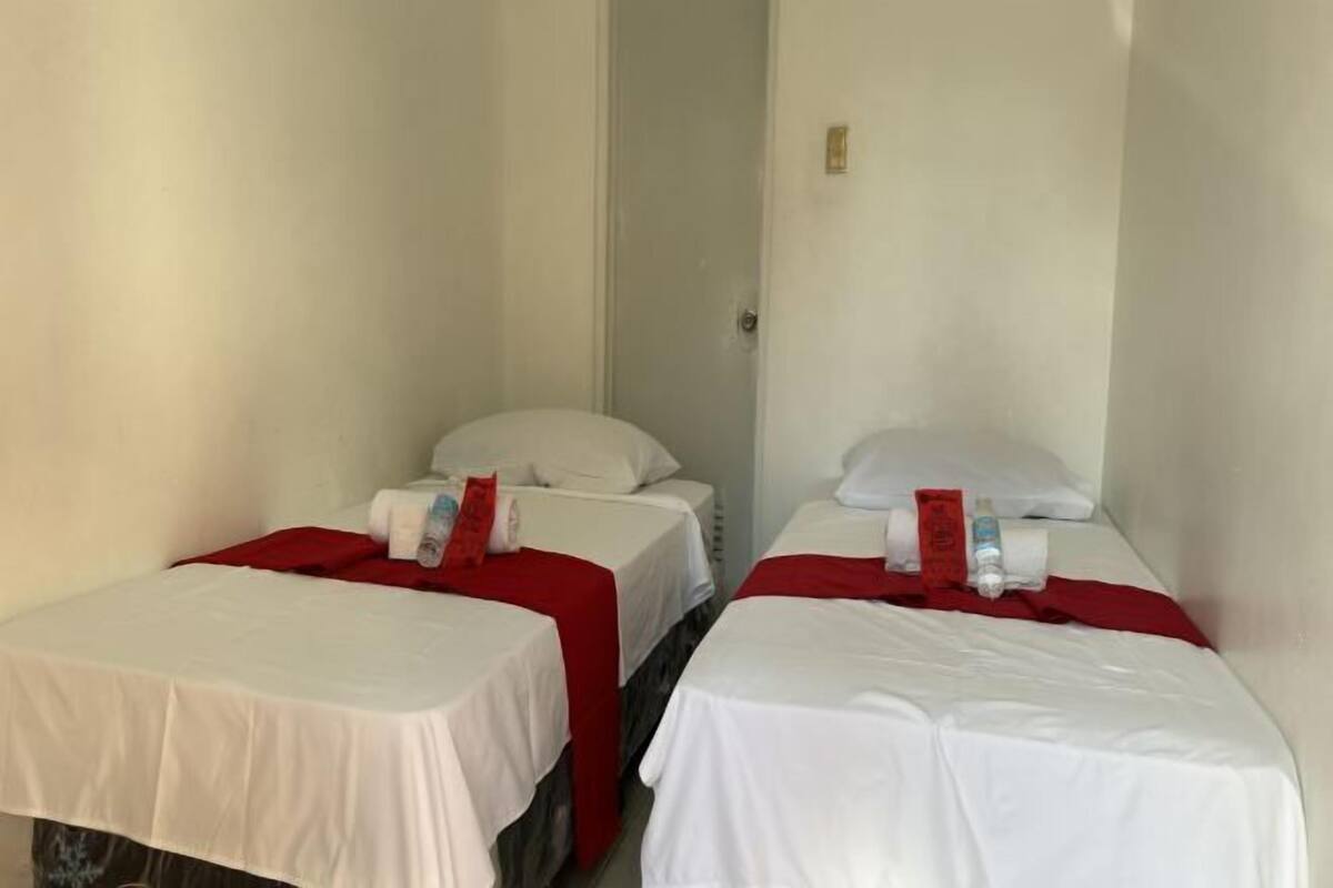 Deux lits simples, avec du linge de lit blanc et des couvre-lits rouges, dans une chambre étroite aux murs blancs.