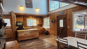 L’intérieur d’un chalet rustique comprend une table à manger en bois, un coin cuisine avec un îlot central et une grande fenêtre qui laisse entrer la lumière naturelle.