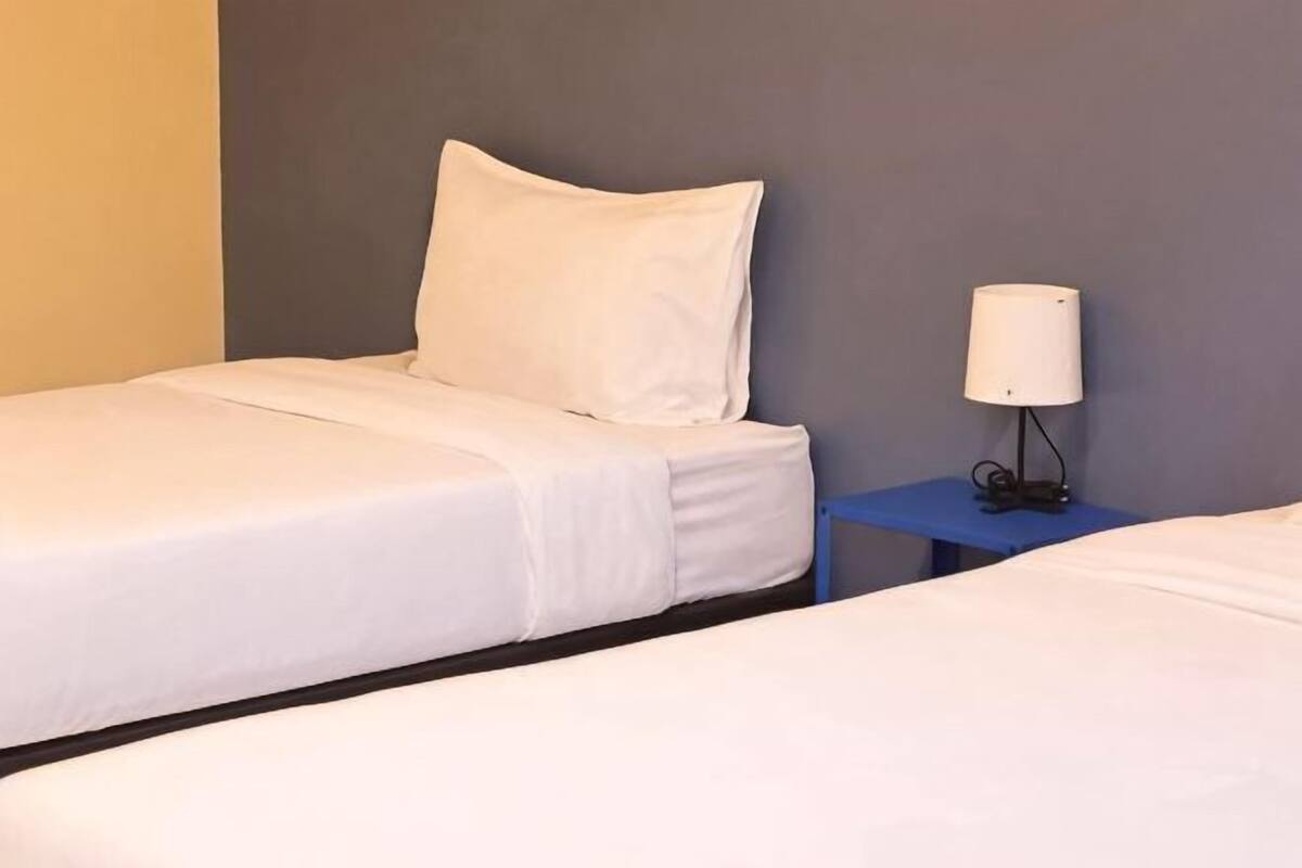 A hotel room with two beds, a nightstand with a lamp, and a plain wall.