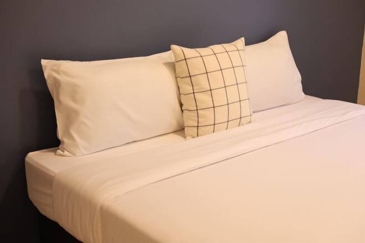 A bed with two pillows, one plain and one with a grid pattern, against a dark wall.