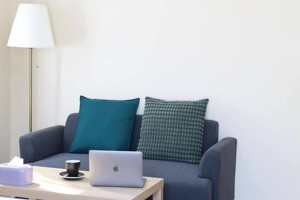A modern living room with a grey sofa, teal and green patterned cushions, a wooden side table with a laptop, a cup, and a tissue box.