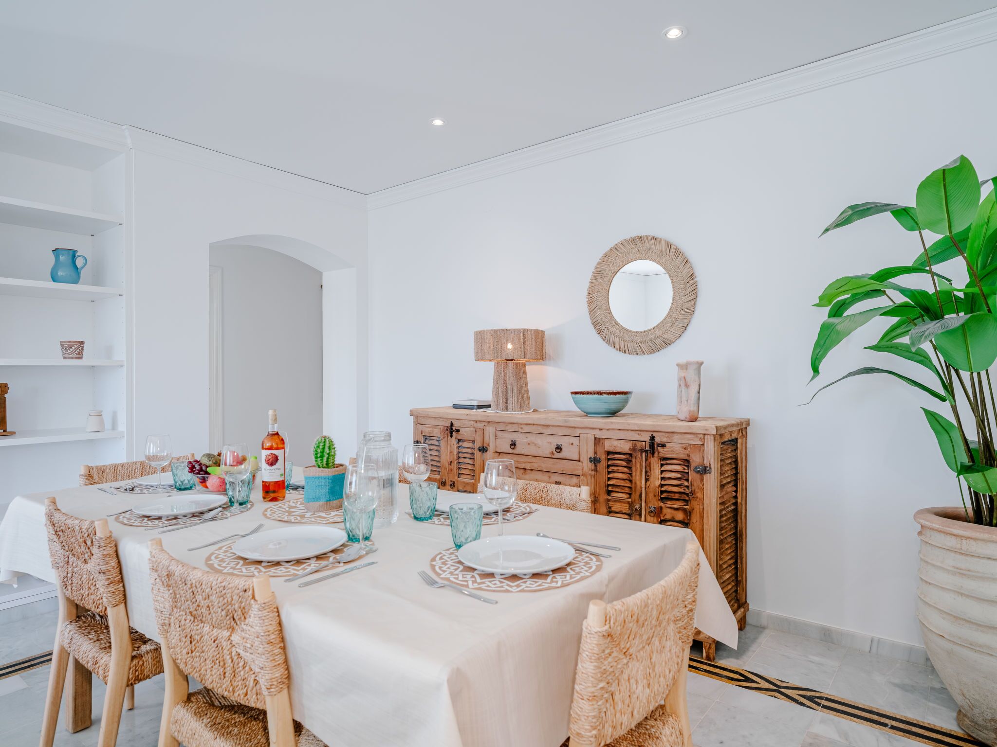 A dining area with a table set for four, surrounded by wicker chairs. A wooden cabinet with a mirror and decorative items is against the wall.