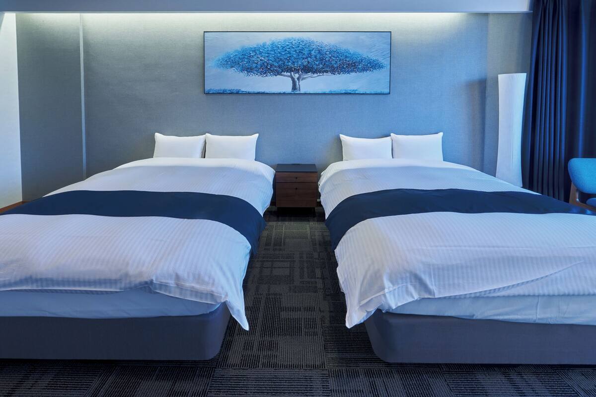 Une chambre d’hôtel avec deux lits, une table de chevet, un tableau représentant un arbre et une chaise.