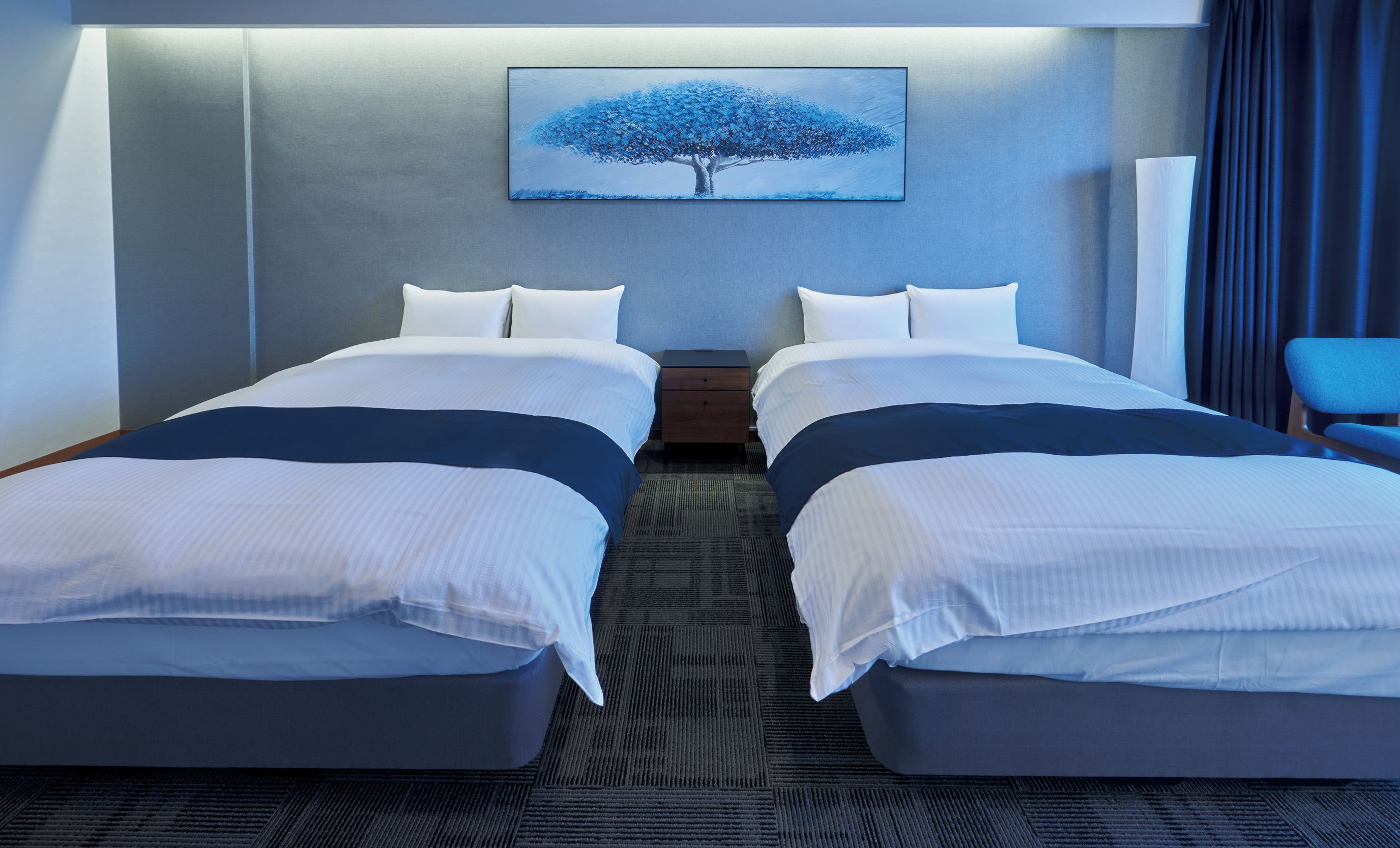 A hotel room with two beds, a nightstand, a painting of a tree, and a chair.