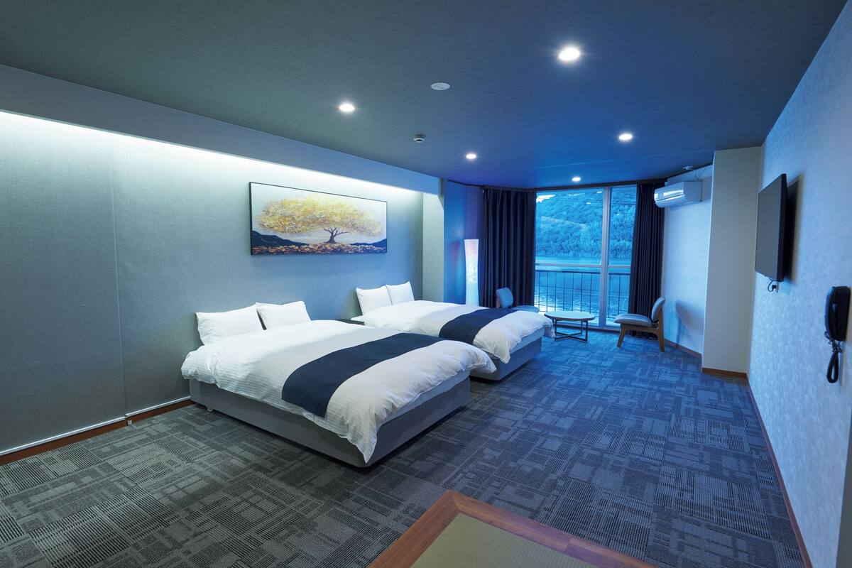 Une chambre d’hôtel avec deux lits, une grande fenêtre donnant sur un beau paysage, un tableau accroché au mur et un téléviseur à écran plat fixé au mur.