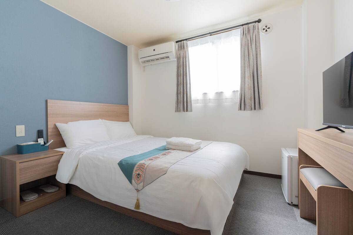 A hotel room with a bed, a desk with a TV, an air conditioner, and a window with curtains.