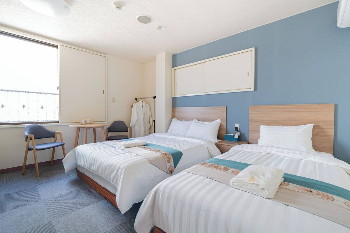 A hotel room with two beds, a desk, a chair, and a window.