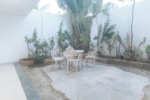 An outdoor seating area with white chairs and a table, surrounded by various plants and a large tree. - RedDoorz @ EBM One Building (Pavia)
