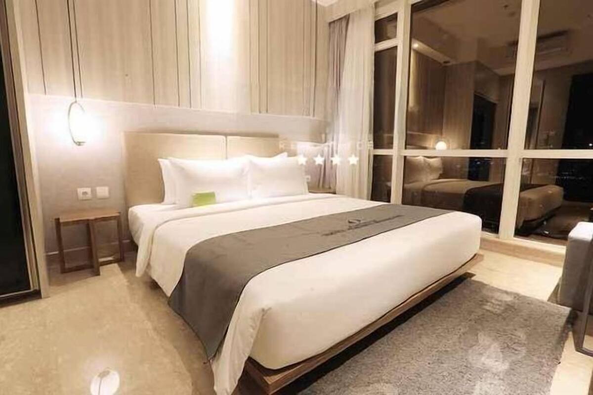 A modern hotel room with a large bed, a nightstand, a small table, and a large window offering a view of the city at night.