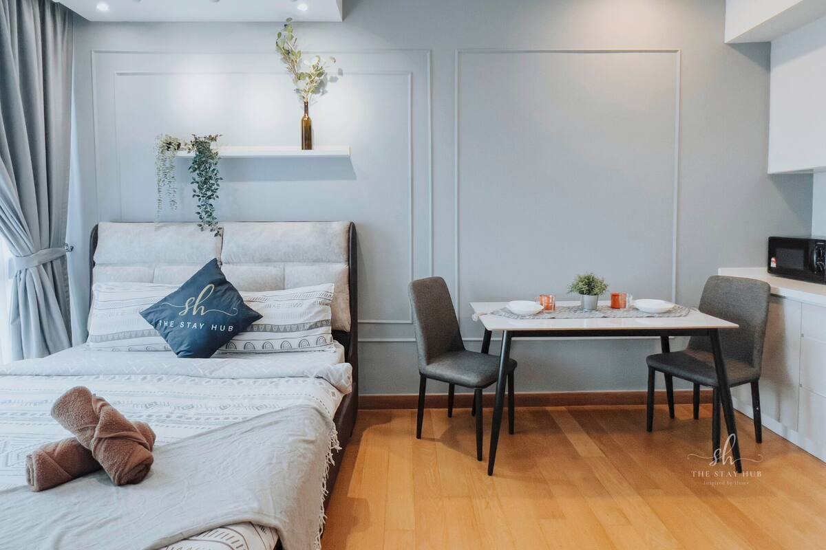 A modern hotel room with a bed, a small dining table with chairs, a microwave, and a potted plant.
