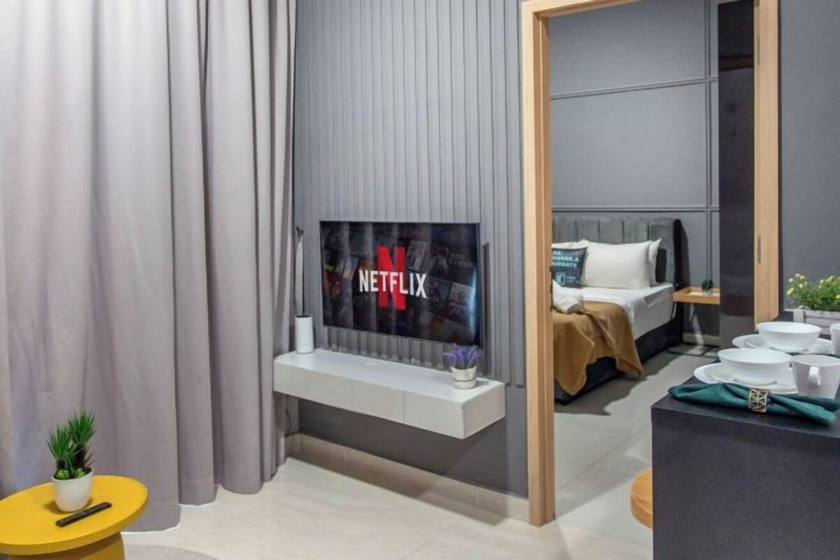 Quarto de hotel moderno com uma televisão de tela plana grande fixada na parede, cama com cabeceira branca, mesa de cabeceira com abajur e uma pequena mesa redonda com uma planta.