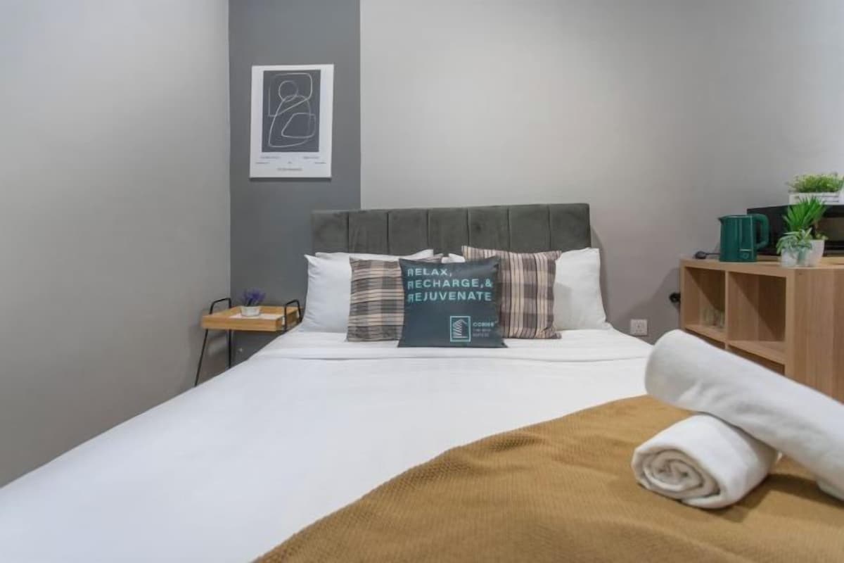 Cama bem arrumada com manta marrom e almofada com a inscrição 'RELAX, RECHARGE, & REJUVENATE'.