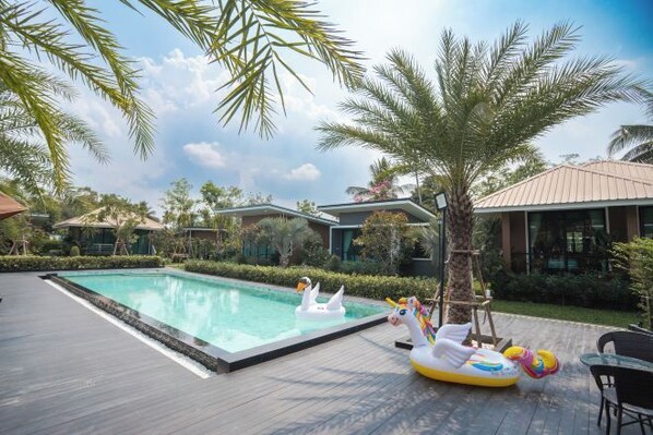 Piscina com brinquedos flutuantes de cisne e unicórnio, cercada por palmeiras e edifícios modernos.