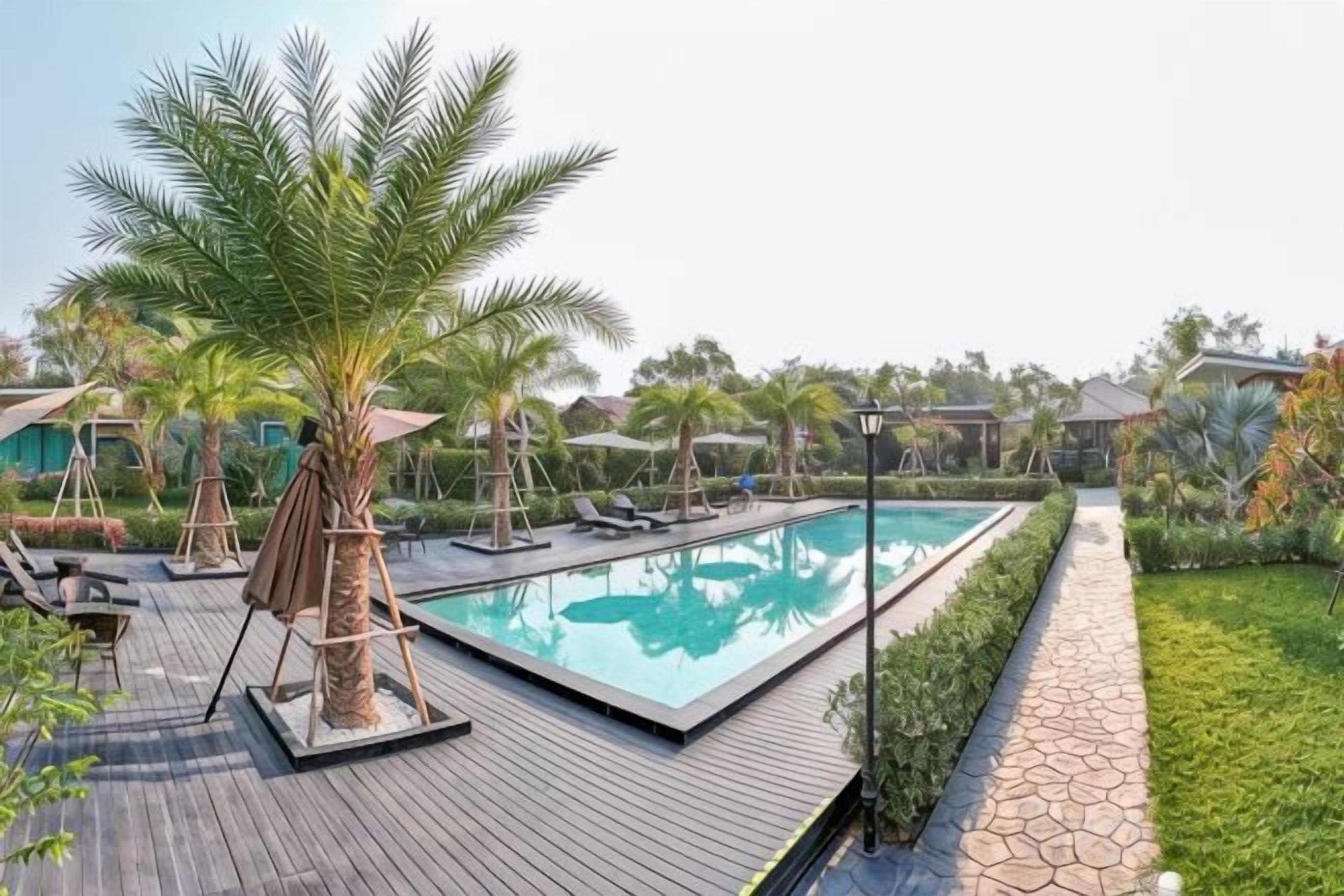 Una piscina con sillas de descanso y palmeras, rodeada por una terraza de madera y un sendero de piedra.