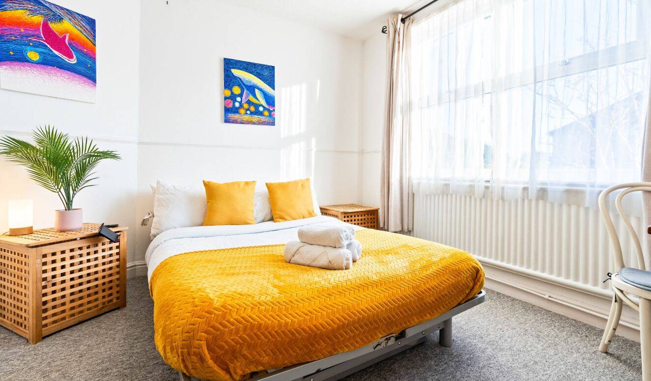 A neatly made bed with a yellow blanket, two pillows, and folded towels. There is a potted plant, a small wooden side table, and a chair.