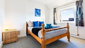 Een slaapkamer met een houten bed, blauw beddengoed, een radiator, een raam met gordijnen en een klein nachtkastje.
