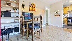 Een moderne woonkamer met een houten tv-meubel, een Netflix logo, een boekenkast, en een keuken in de achtergrond.
