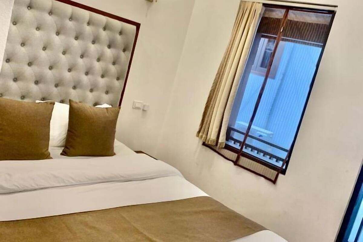 A bedroom with a tufted headboard, a large window with curtains, and a bed with white and brown bedding.