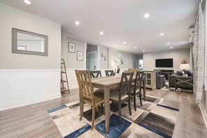 A dining area with a wooden table and chairs, a kitchen in the background, and a living area with a TV.