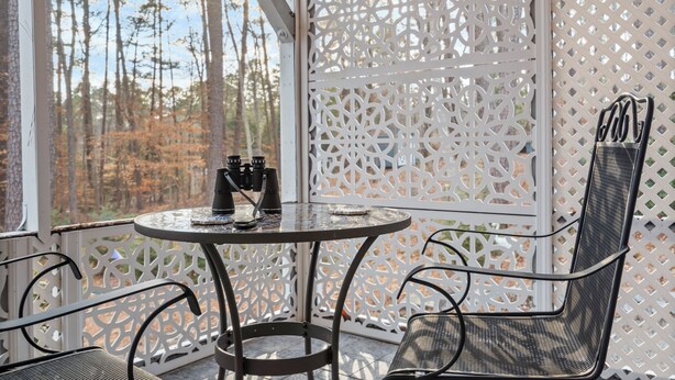 Un espacio al aire libre techado con mesa y sillas, vista a árboles y un panel de celosía blanca.