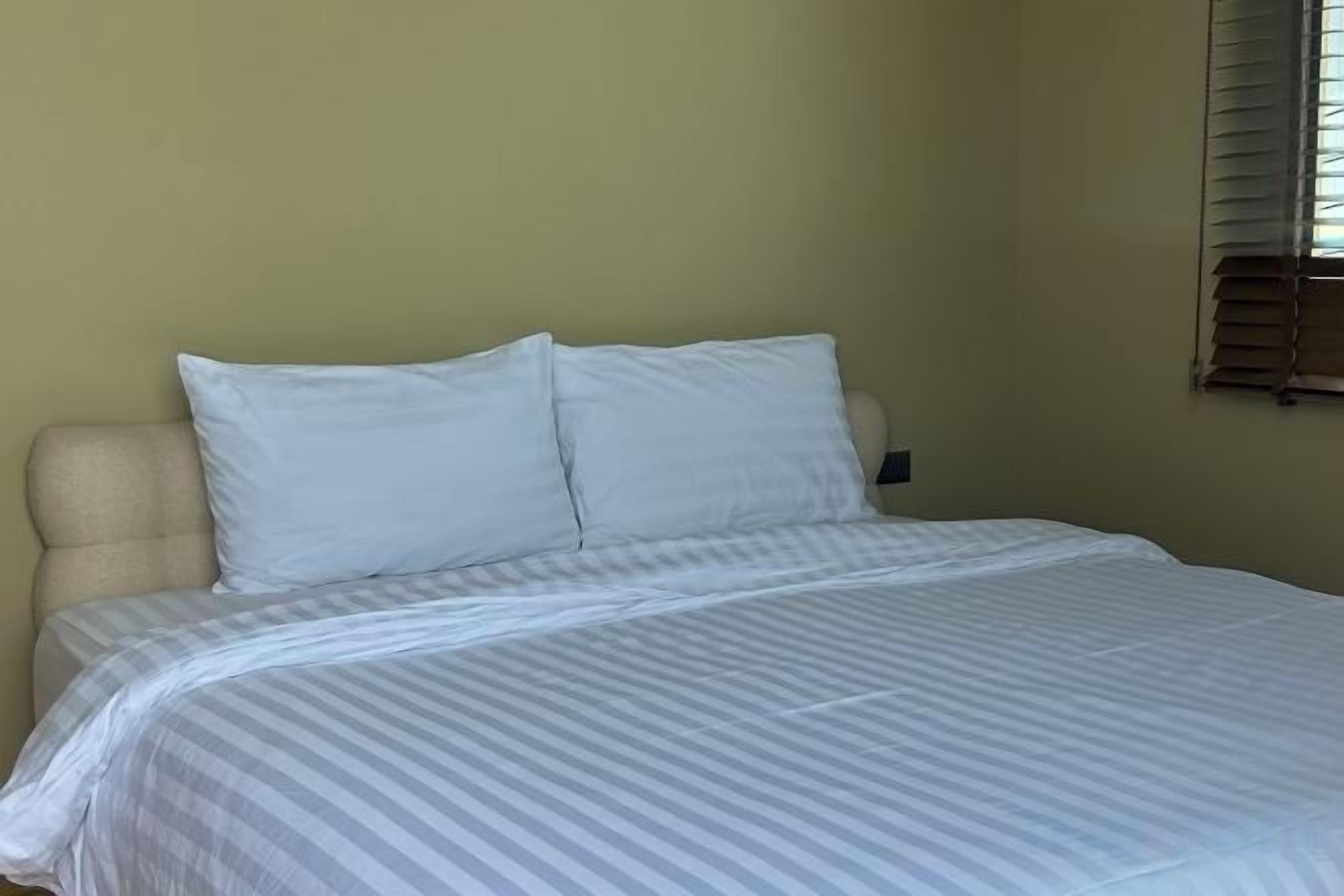 A bed with a striped bedspread and two pillows in a room with a plain wall and a window with blinds.