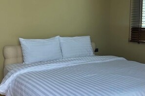 Una cama con una colcha a rayas y dos almohadas en una habitación con una pared lisa y un ventanal con persianas.