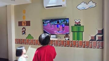 Una habitación con una televisión mostrando un videojuego, un aire acondicionado instalado en la pared y un diseño temático de Mario en la pared.