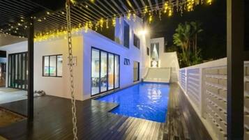A swimming pool with a wooden deck, a covered area with string lights, and a white building with large windows.