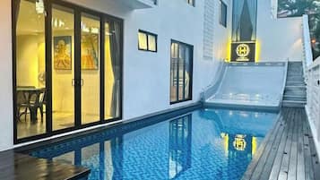 A swimming pool with a wooden deck, a glass door entrance, and a small outdoor seating area.