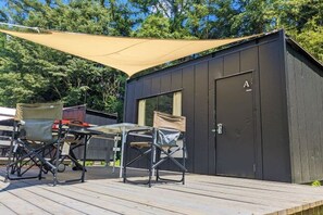 A small, dark-colored building with a door marked 'A' and a window. There is a wooden deck in front with two folding chairs and a table. - Bulls Camp -Guesthouse with pets at the foot of World Heritage Mt.Fuji- (Narusawa)