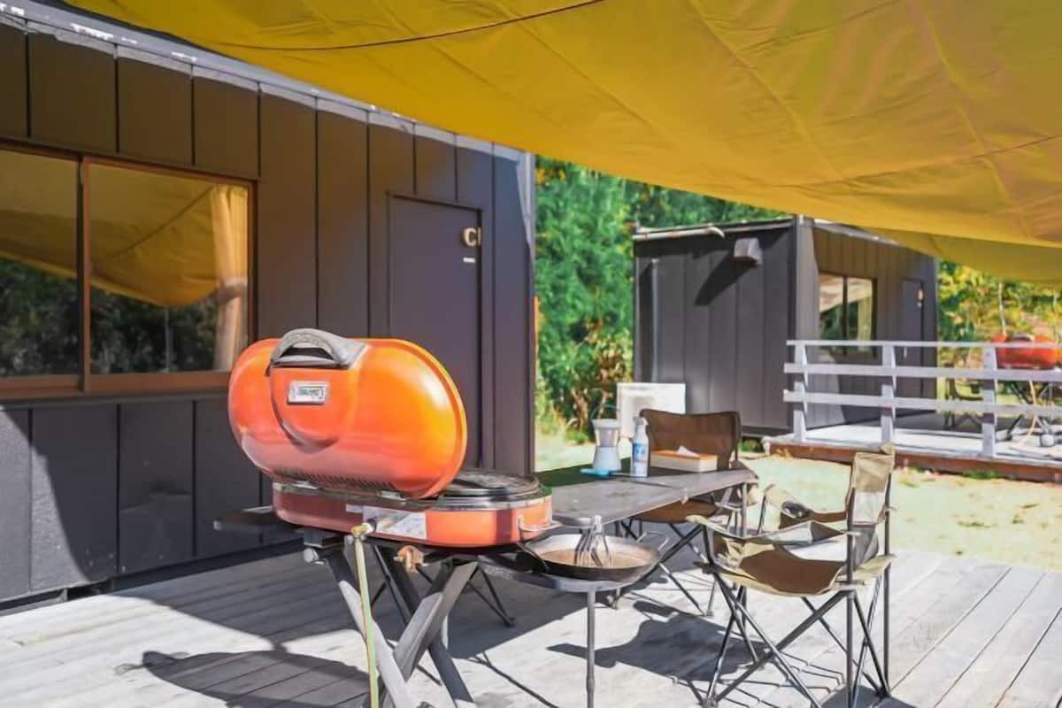 A barbecue grill on a wooden deck with a table and chairs, a yellow canopy, and a small building in the background.