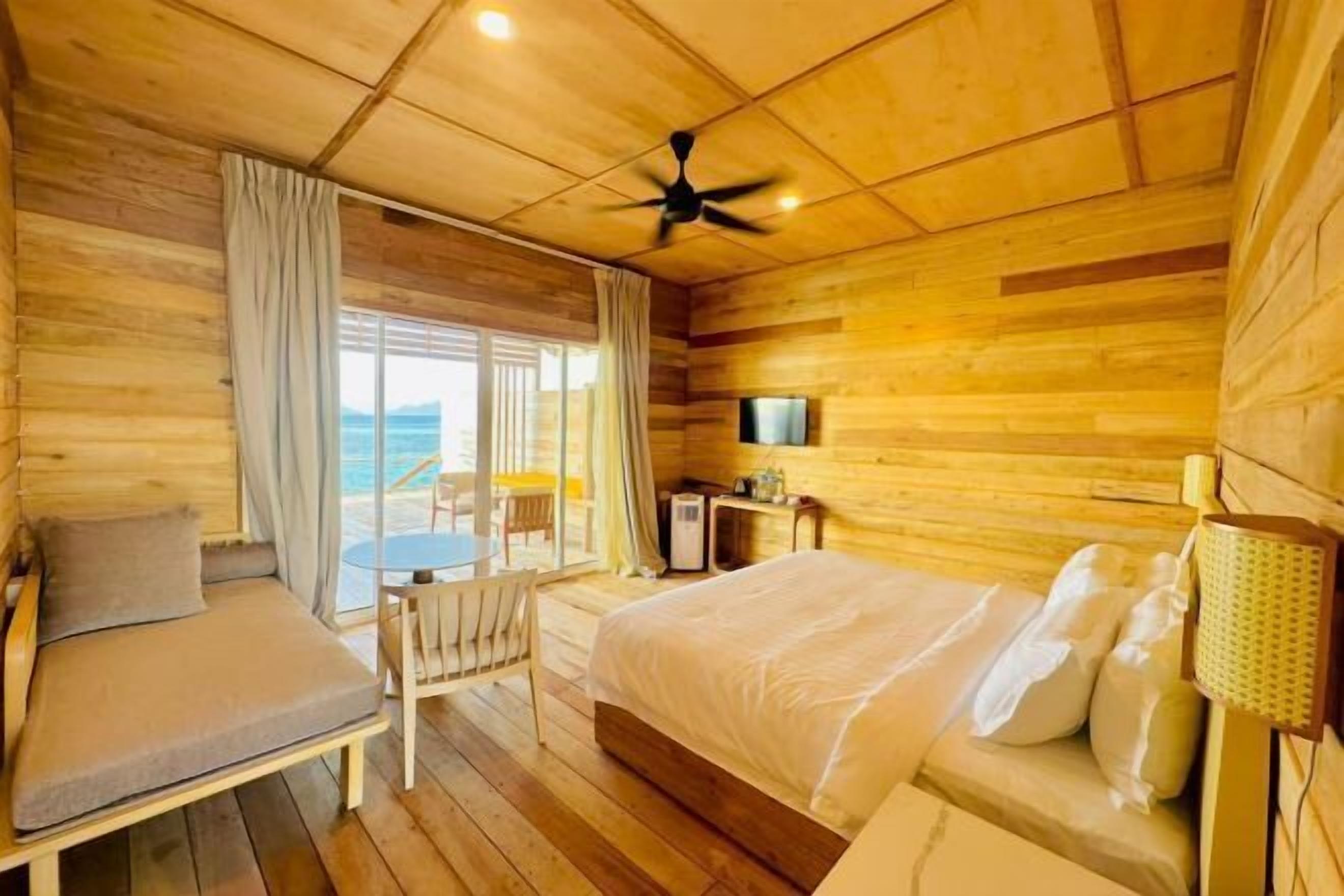 A wooden cabin room with a bed, a chair, a table, a ceiling fan, and a TV.
