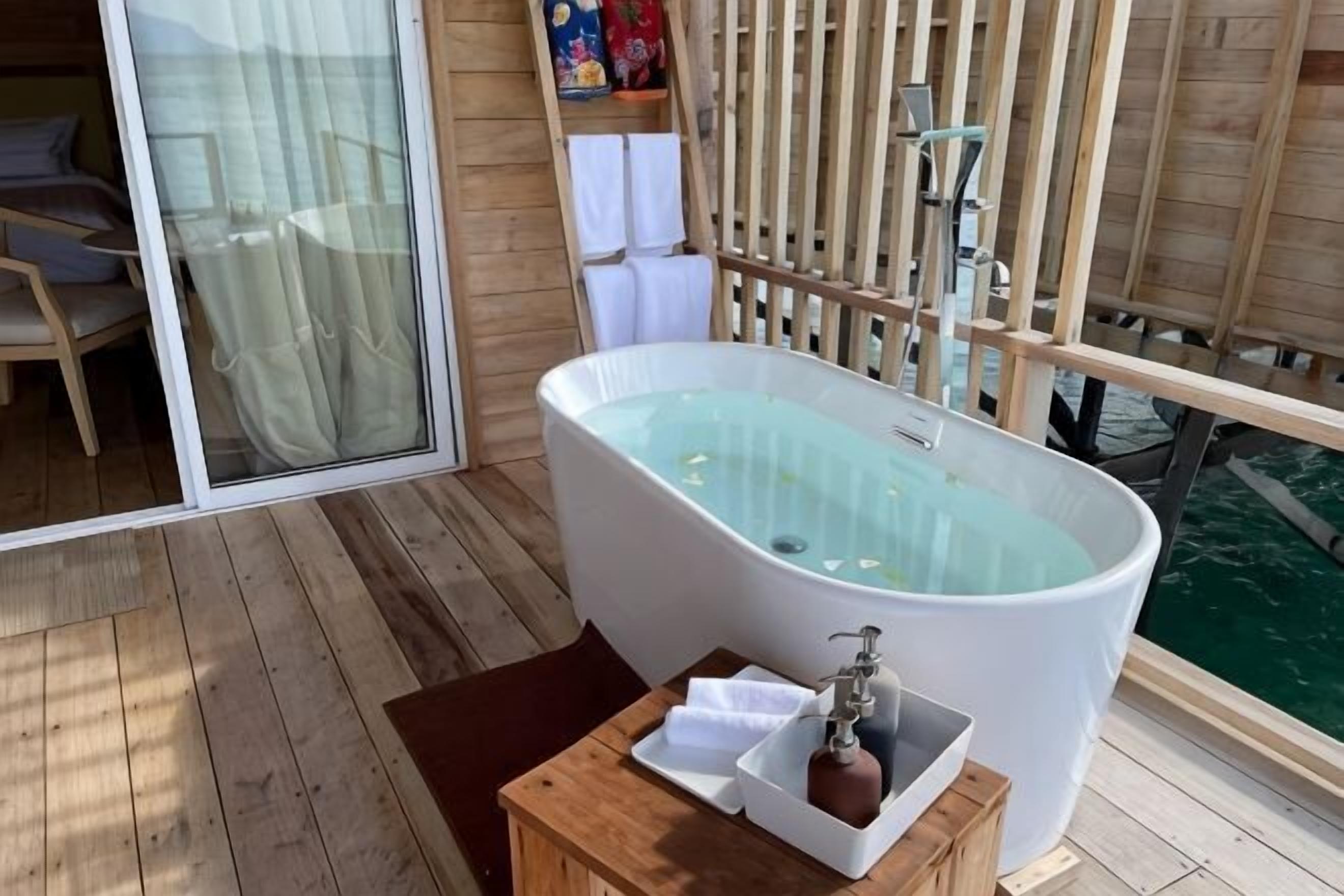 A room with a large bathtub, wooden flooring, a small table with a towel and a bottle, and a view of the water outside.